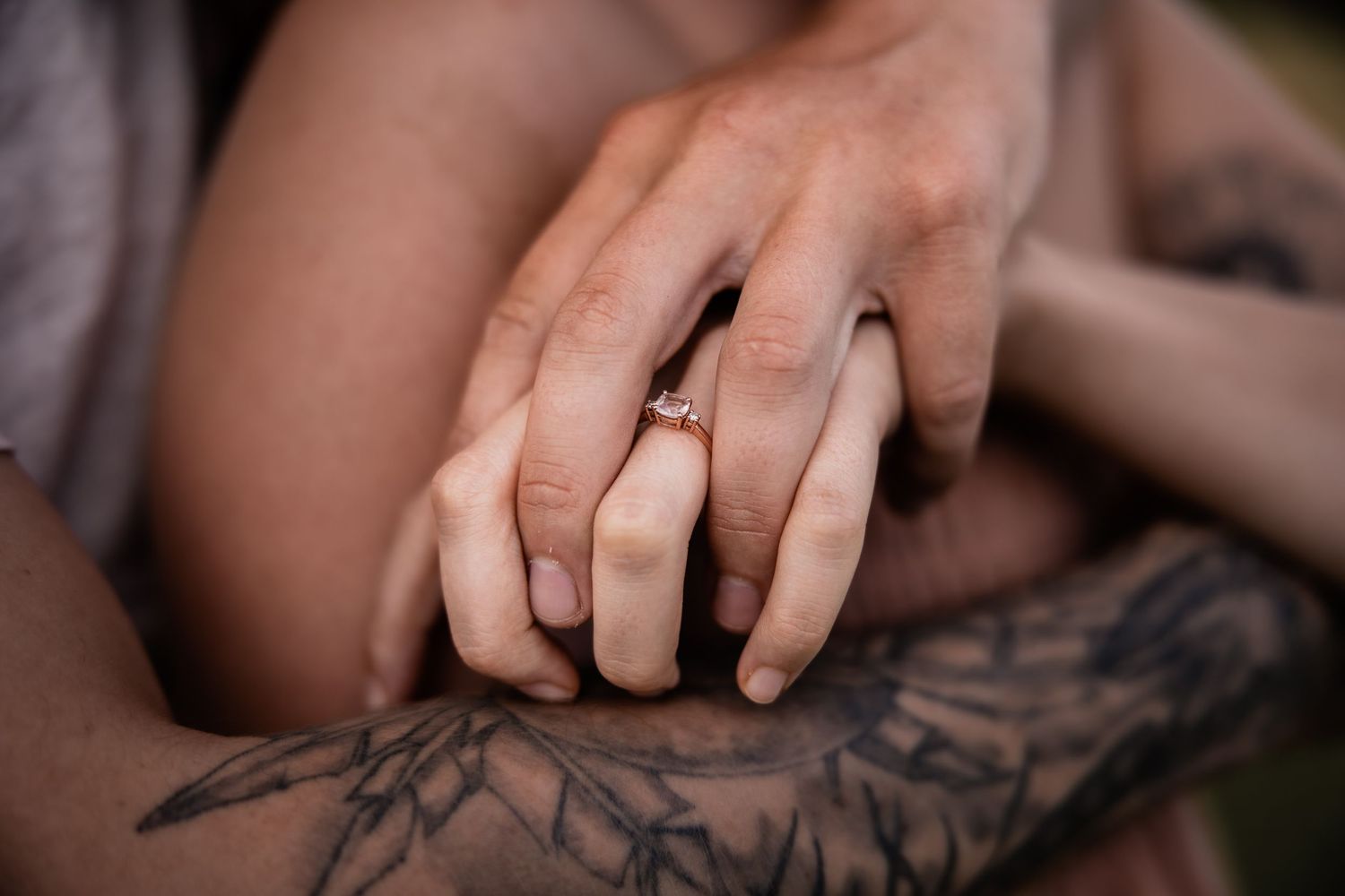 Close-up of intertwined hands with engagement ring and visible tattoo artwork.