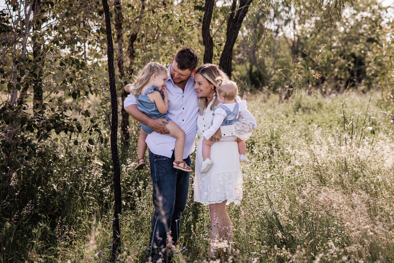 Family stands together in a sunlit meadow surrounded by trees during a casual outdoor photo session.