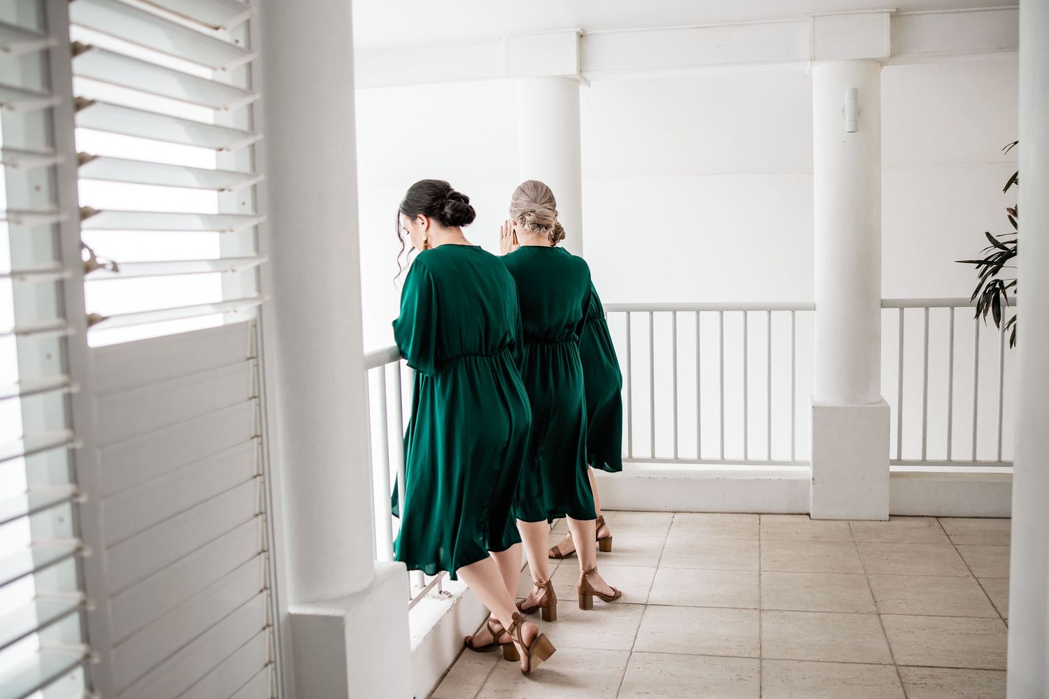Two people in emerald green dresses walking down a bright hallway with white shutters.