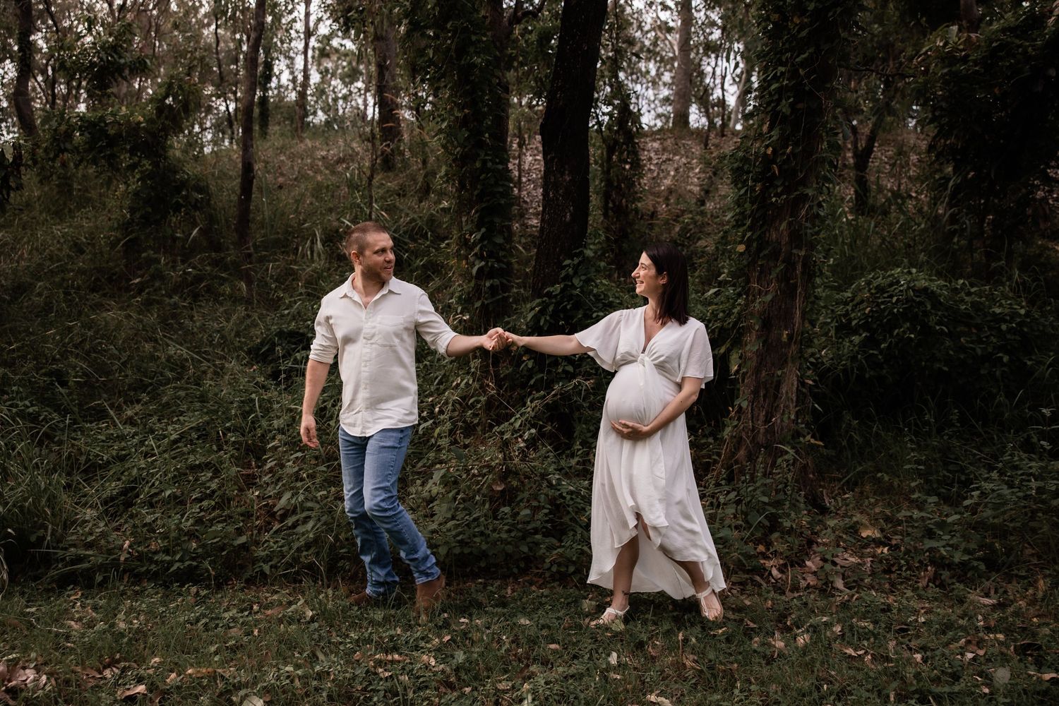 A couple holds hands while walking through a lush green forest setting.