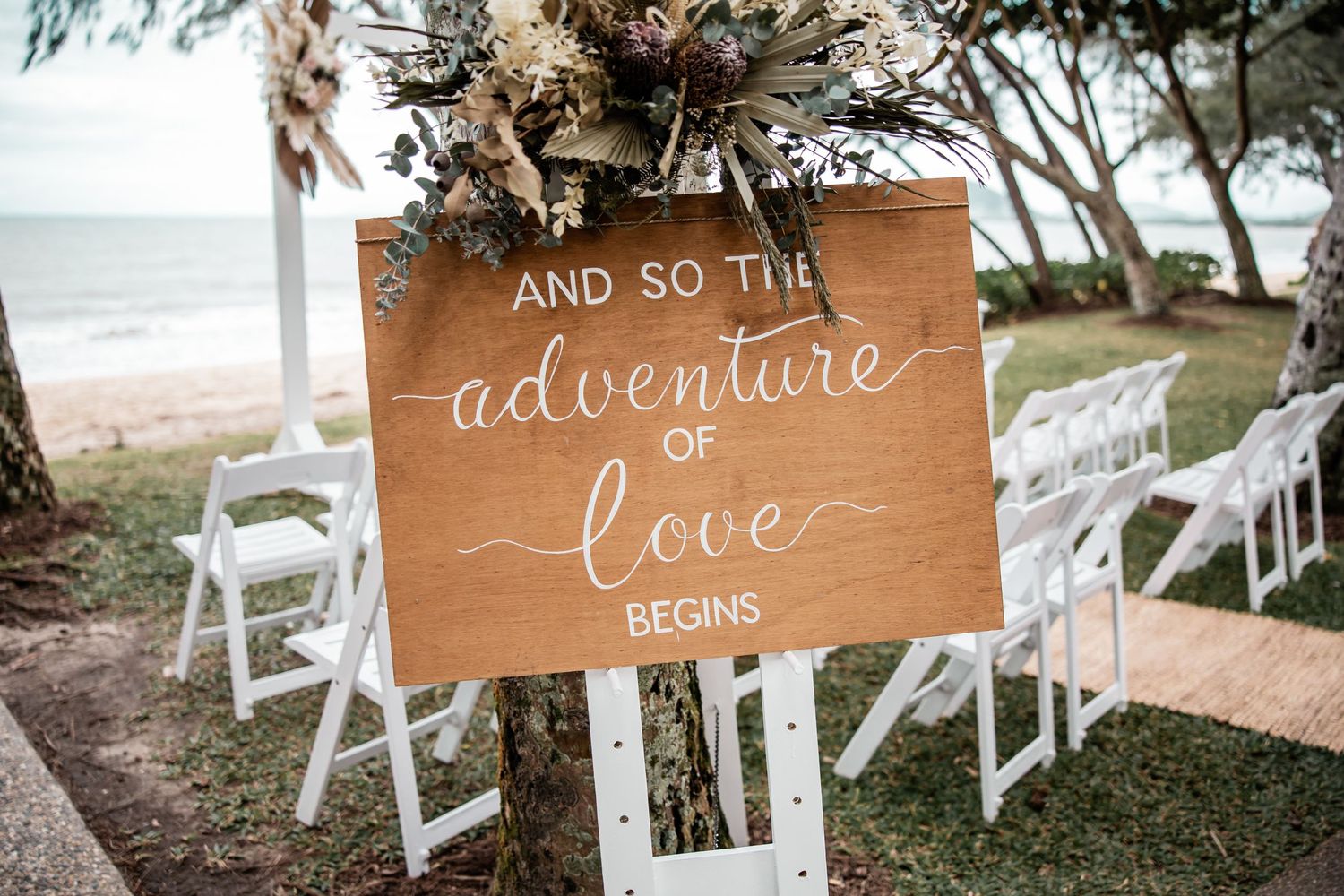 A rustic wooden wedding sign reading 'And so the adventure of love begins' with white chairs on a beach.