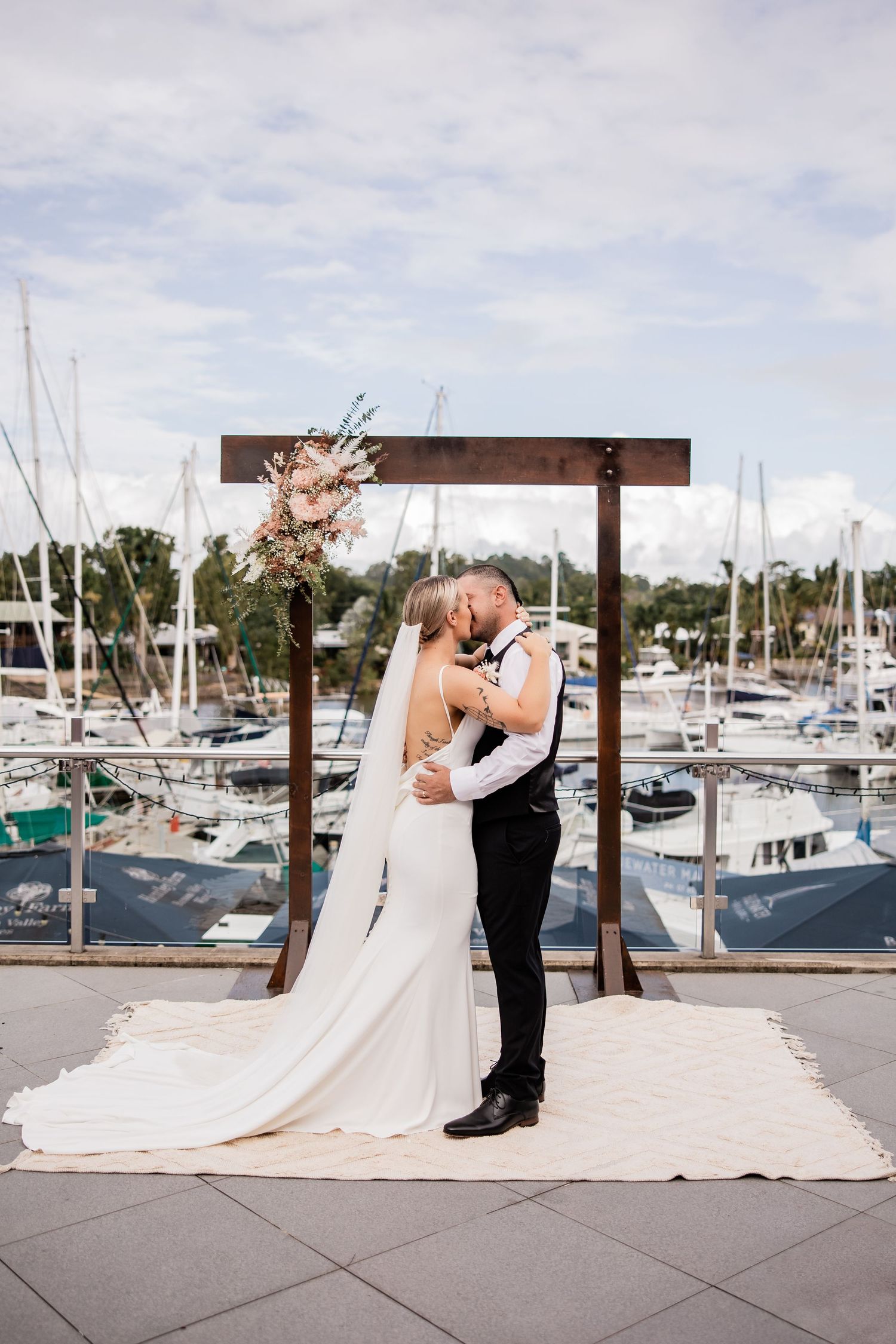 A romantic wedding moment at a marina with boats in the background under a wooden arch.