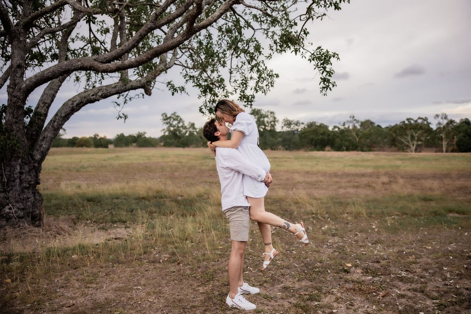 Romantic embrace in a rustic field setting with large tree in background.