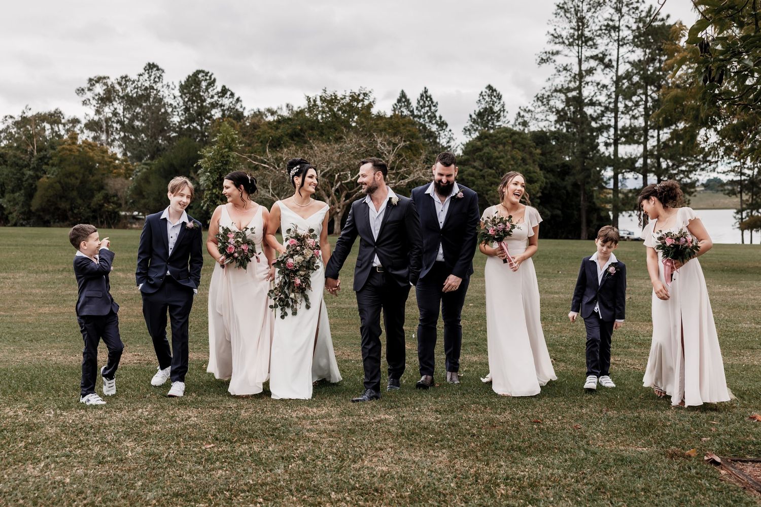Wedding party in formal attire poses on grassy field with bridesmaids in white dresses.