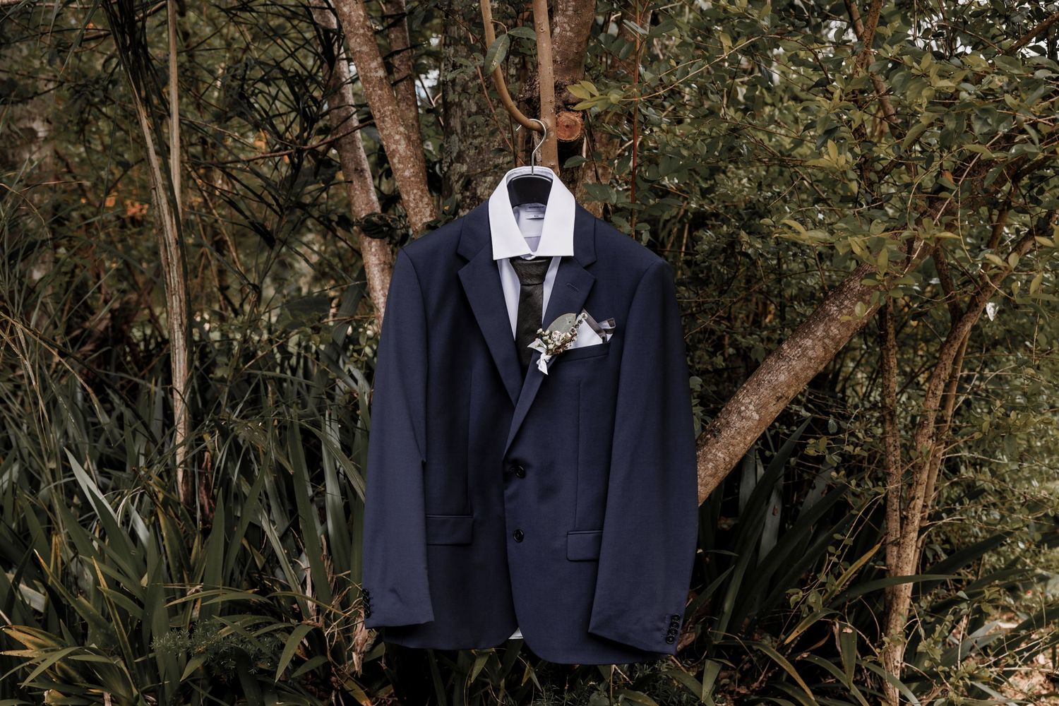 A navy blue groom's suit jacket with boutonniere displayed against natural foliage.