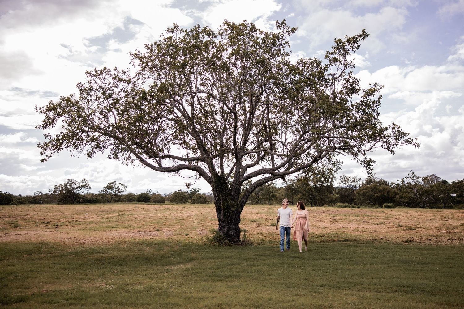 A couple stands beneath a majestic tree in an open field under dramatic clouds.