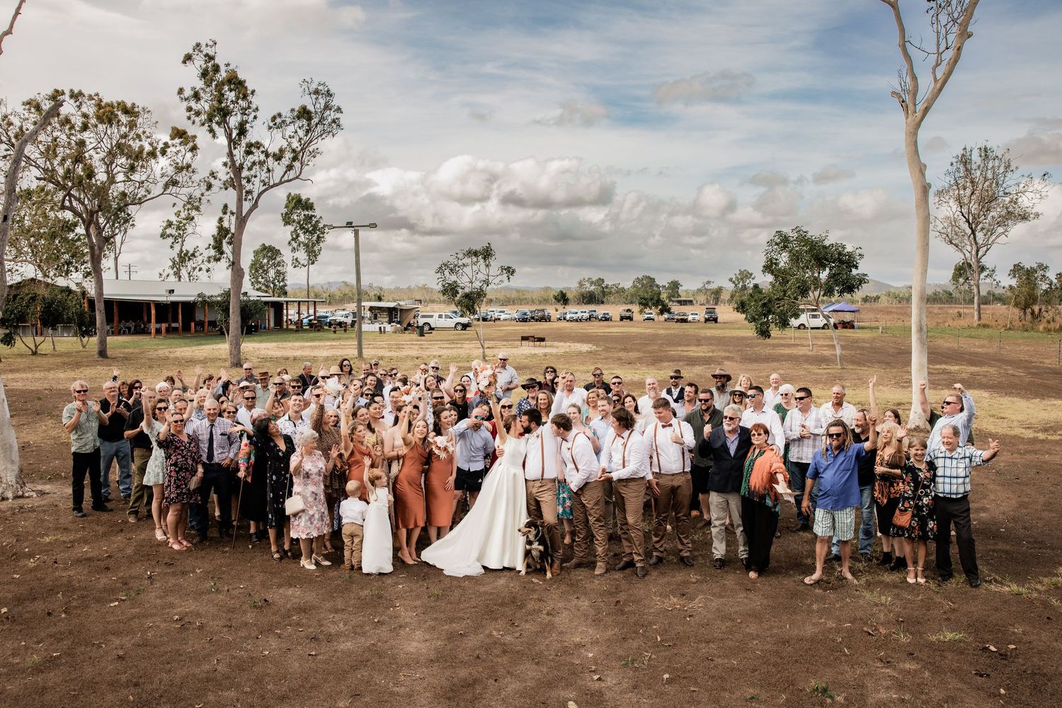 Wedding party and guests pose together in rustic countryside setting.