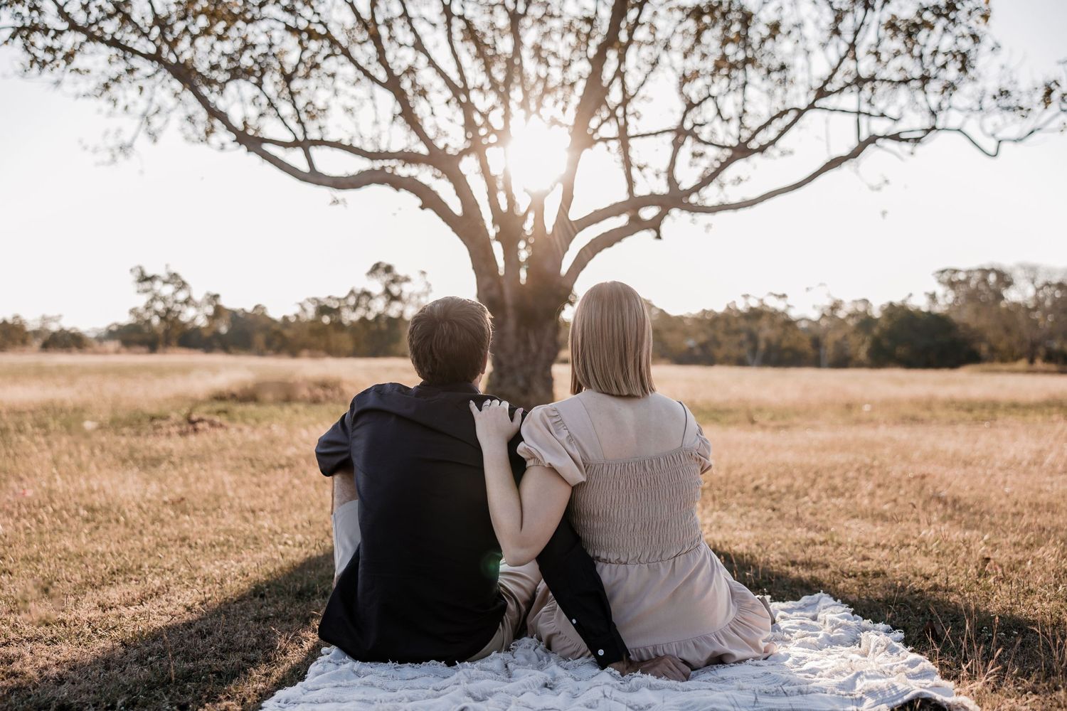 A couple sits on a blanket under a large tree at sunset in a serene countryside setting.