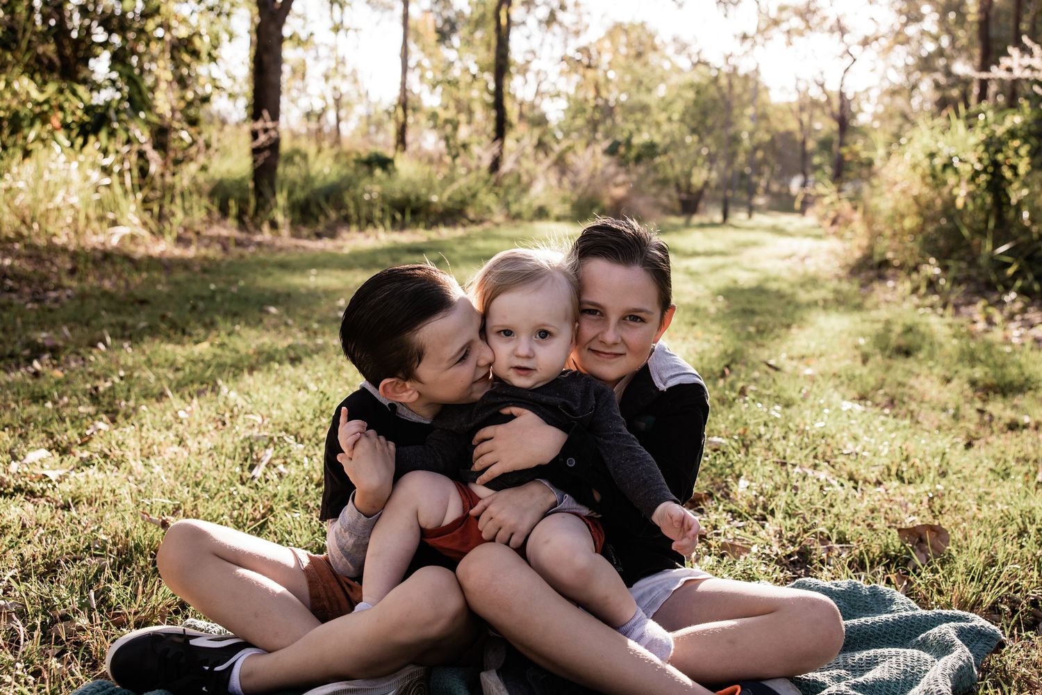 Three children sitting close together on a blanket in a sunlit forest setting.