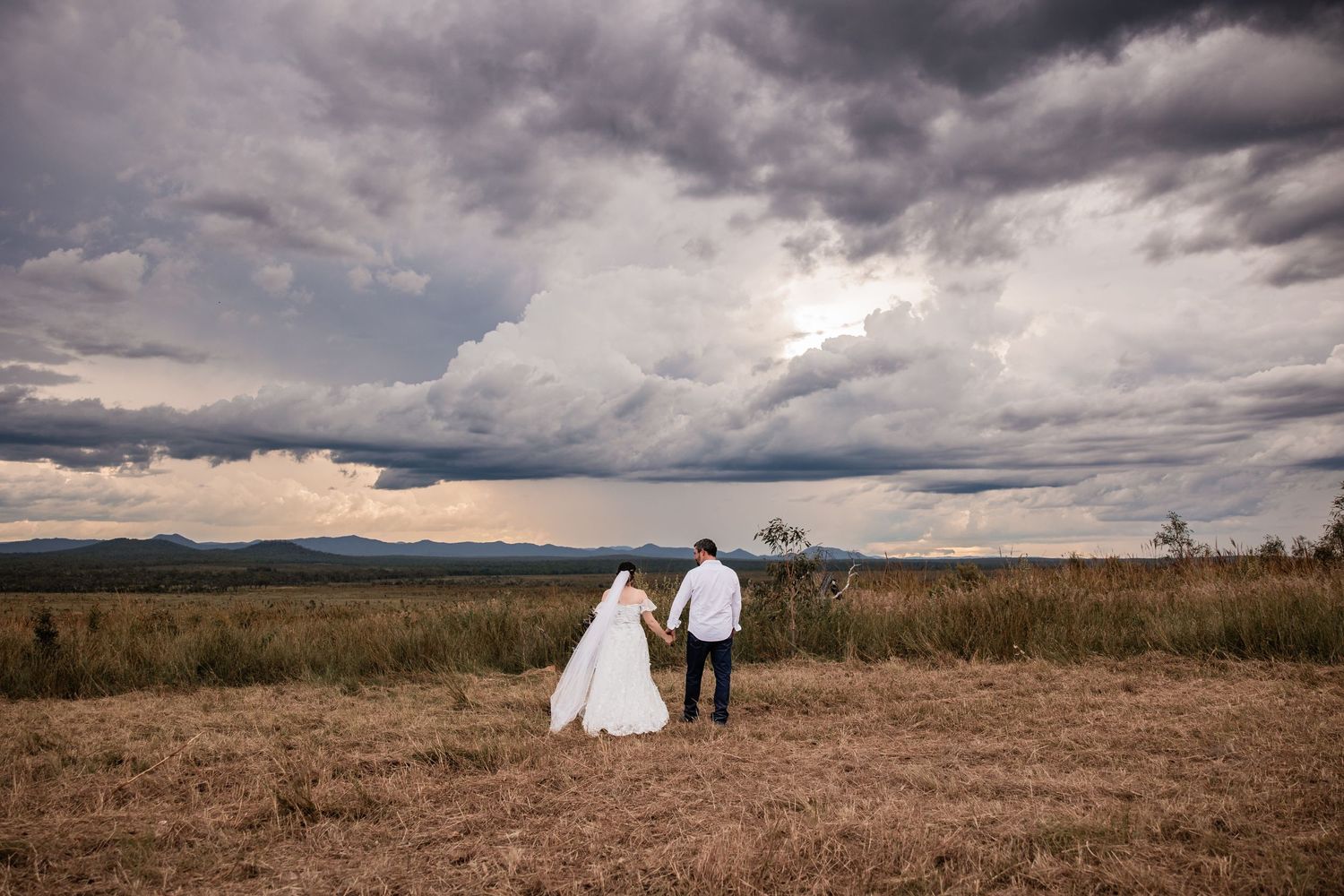 Dramatic storm clouds loom over wedding couple in countryside landscape.