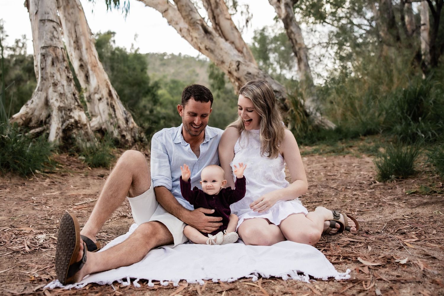 Family relaxes on white blanket under eucalyptus trees during outdoor photo session.