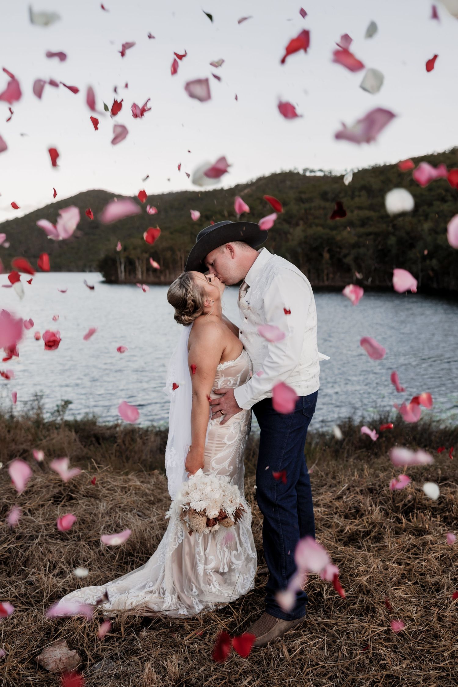 Couple shares romantic moment as rose petals fall around them by lakeside.