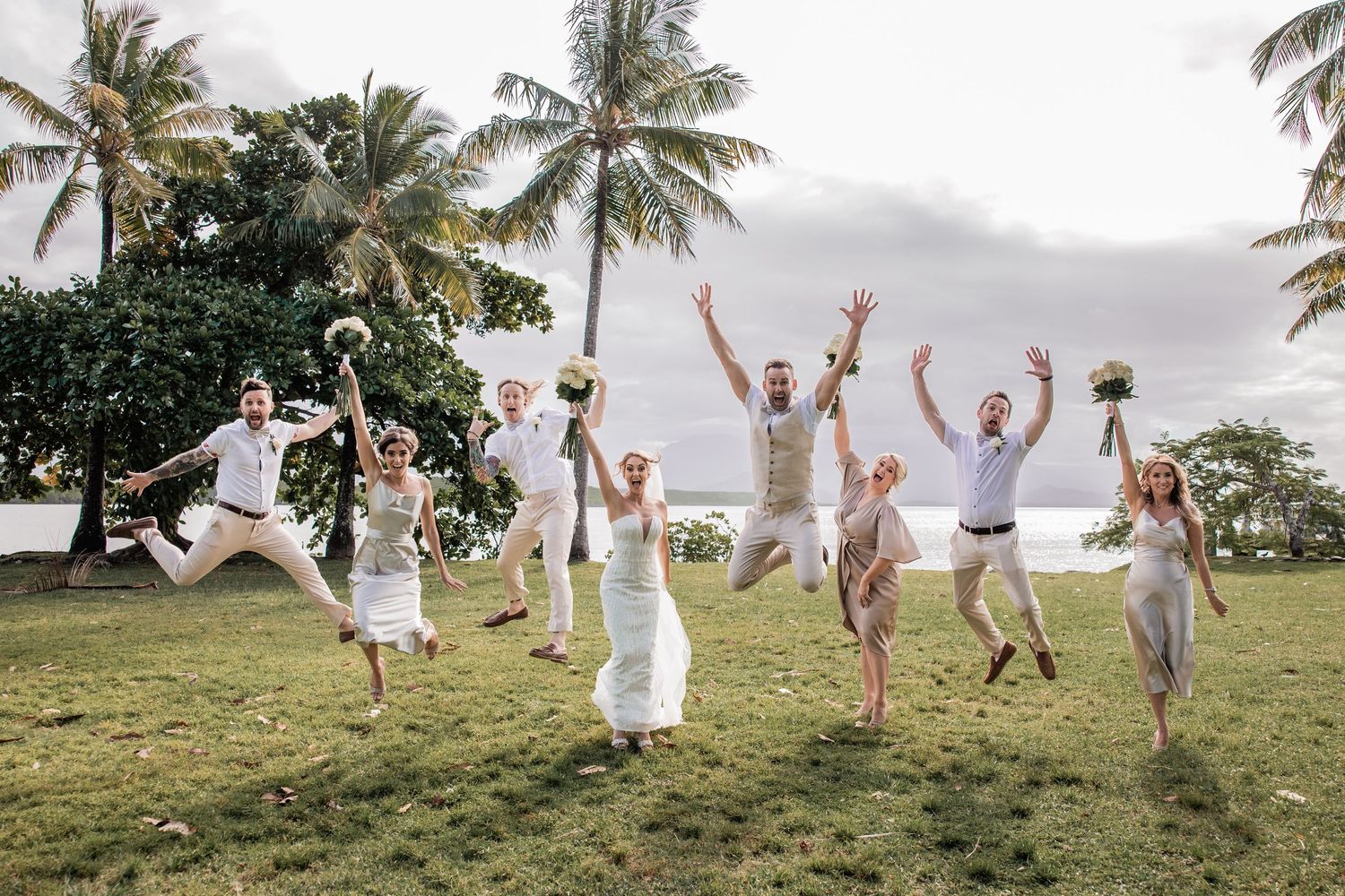 Wedding party jumps joyfully on grass field with palm trees in tropical setting.