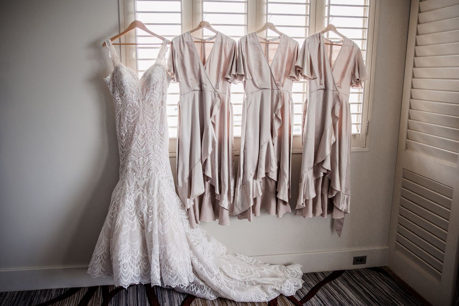 A lace wedding gown hangs alongside blush bridesmaid dresses near a shuttered window.