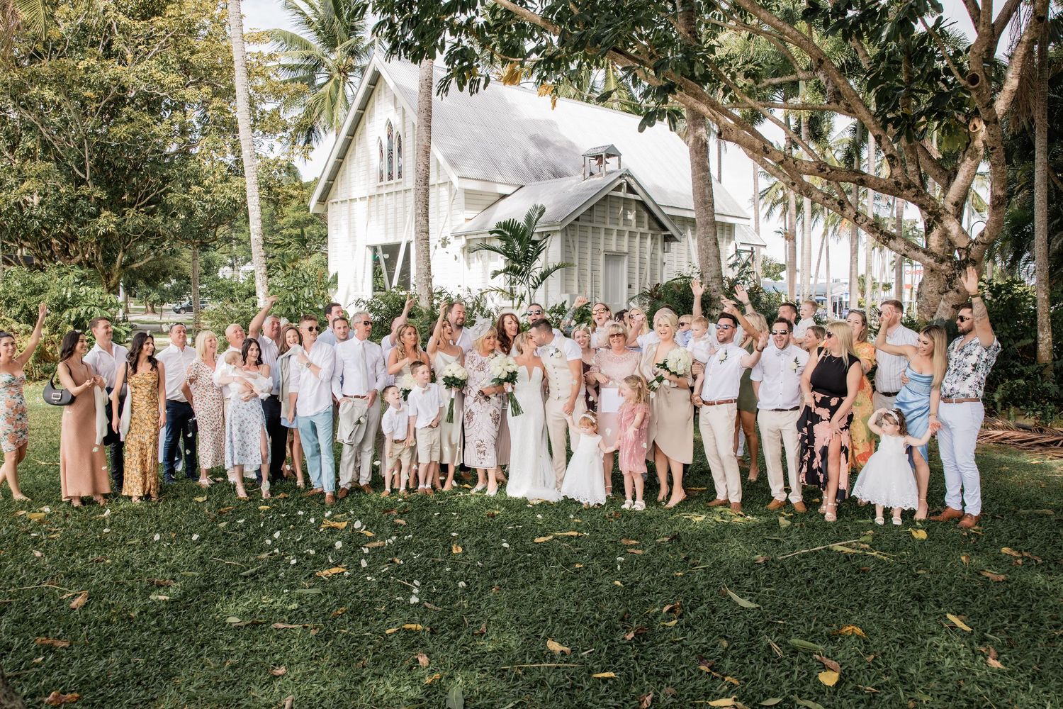 Wedding guests gather on lush green lawn with white chapel and palm trees in background.