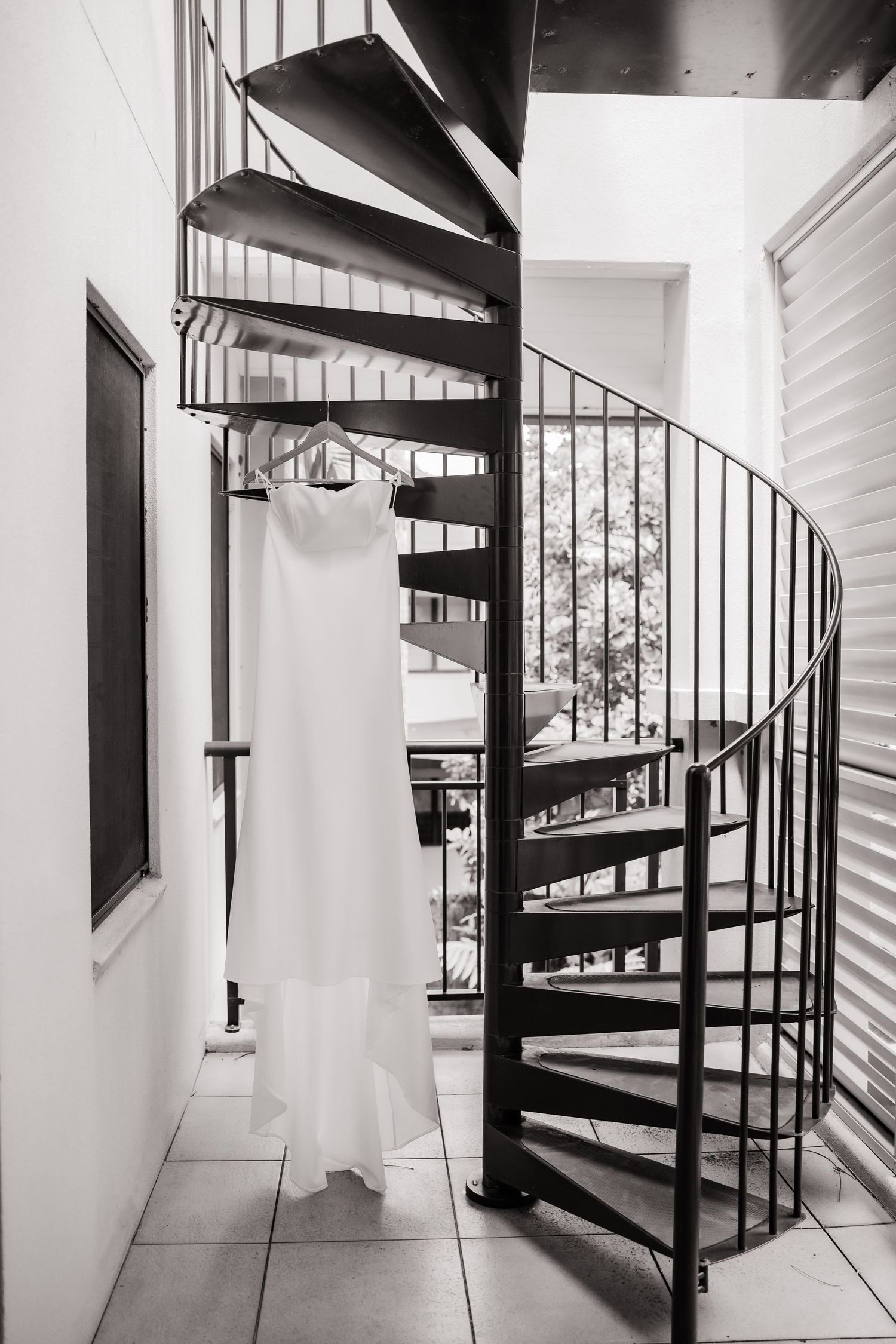 A white wedding dress hangs elegantly on a black spiral staircase in dramatic black and white.