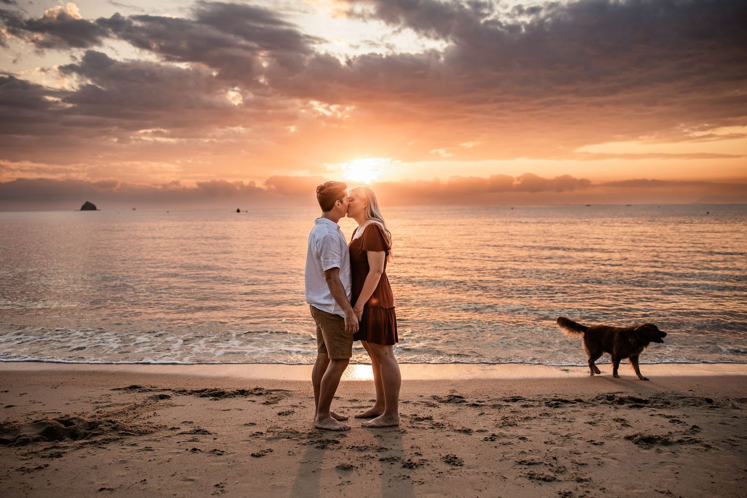 A couple embracing on a beach at sunset with a dog silhouetted against the orange sky.