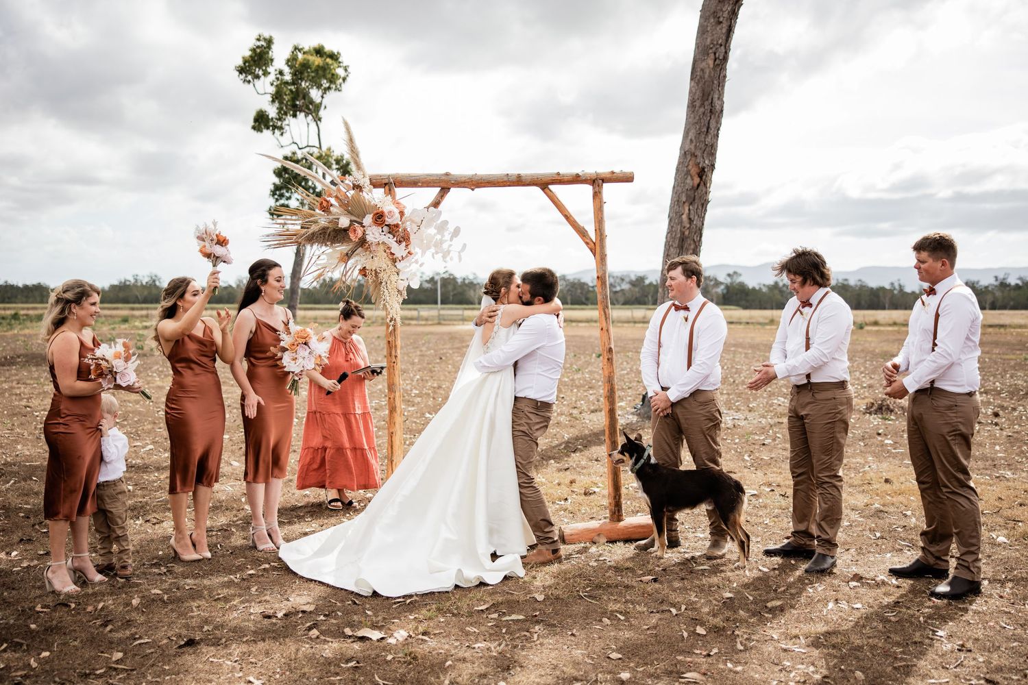 A wedding ceremony under a wooden arch with bridesmaids in rust-colored dresses and groomsmen in suspenders.
