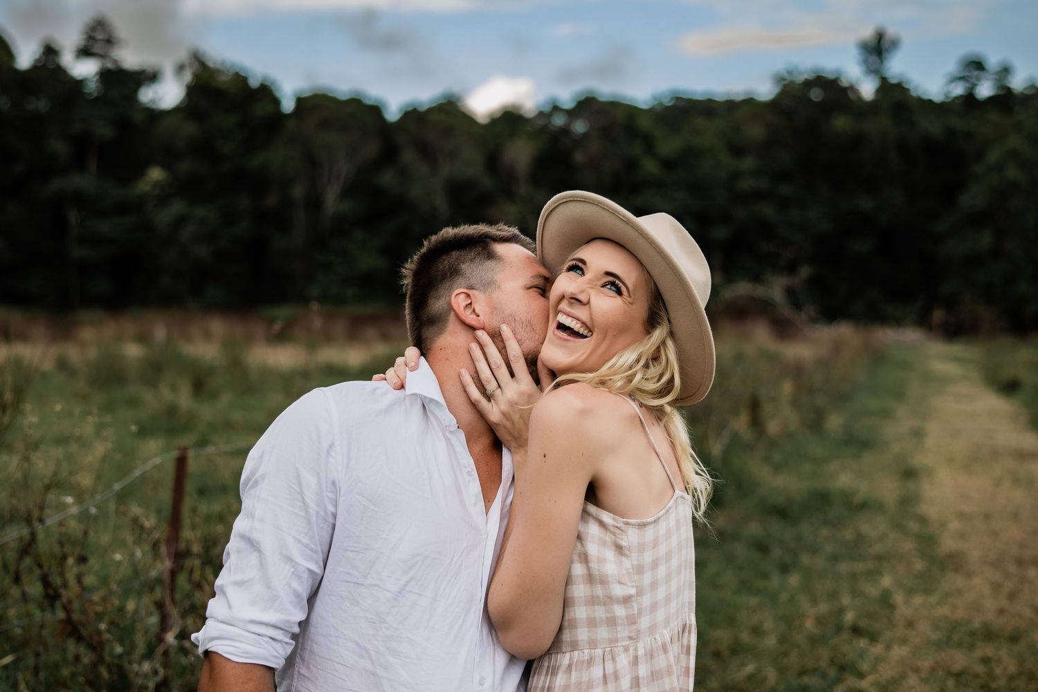 A romantic couple laughing together in a sunny field with a woman wearing a sun hat.