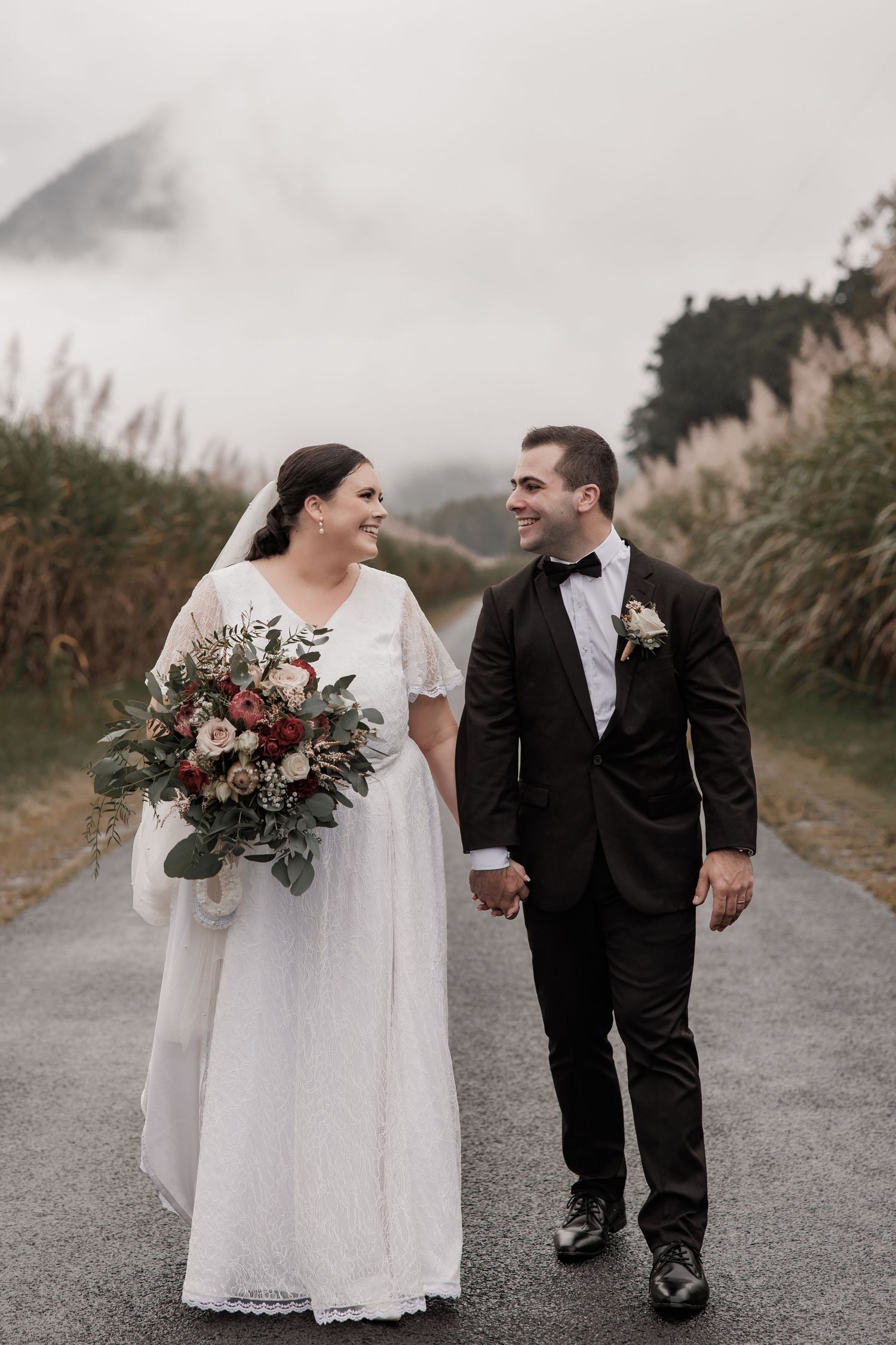 Wedding couple walking together on a foggy path with lush greenery and a beautiful bouquet.