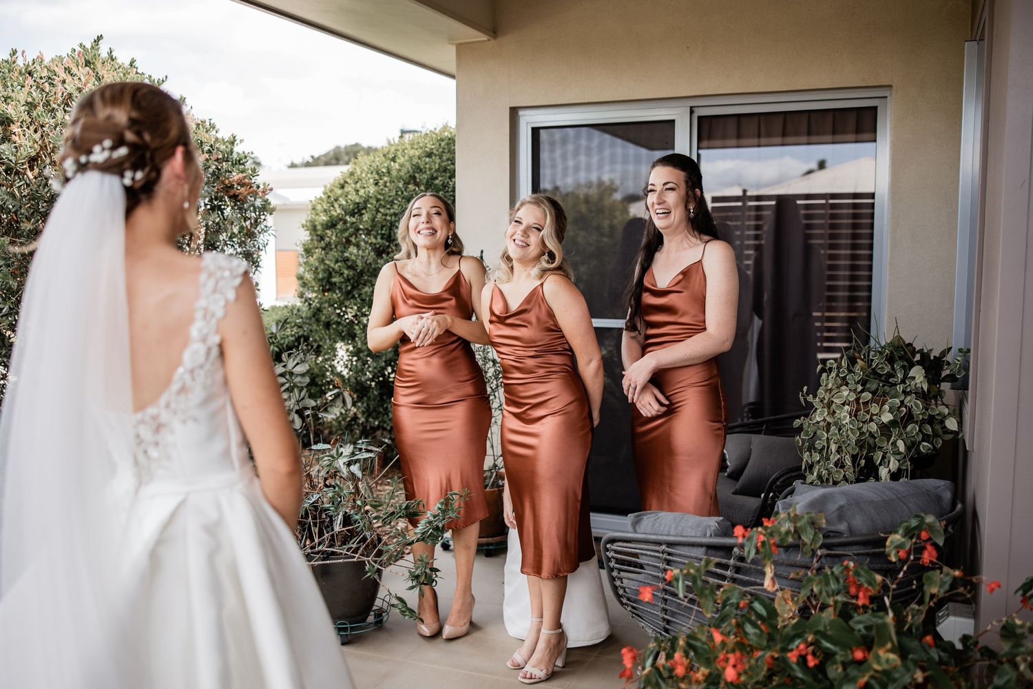 Bridesmaids in silk copper dresses admiring the bride in her white gown on a sunny patio.