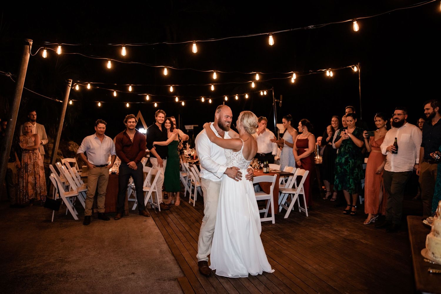 Newlyweds share their first dance on wooden deck under string lights with guests watching.