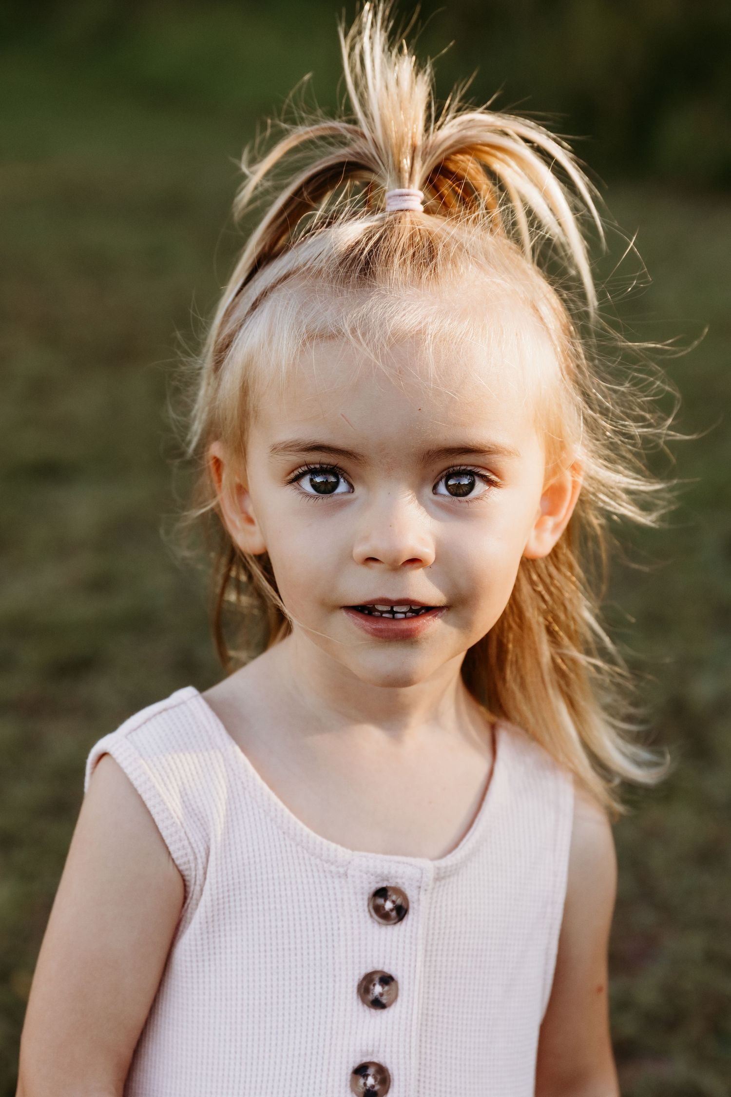 Child in white tank top with blonde hair and sweet smile outdoors.