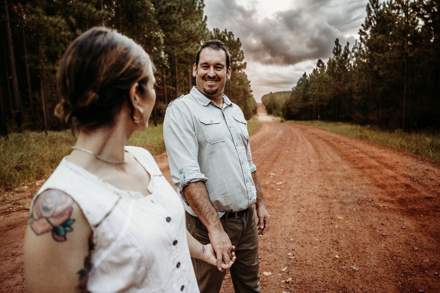 A couple holds hands on a rustic dirt road through a forest under dramatic skies.