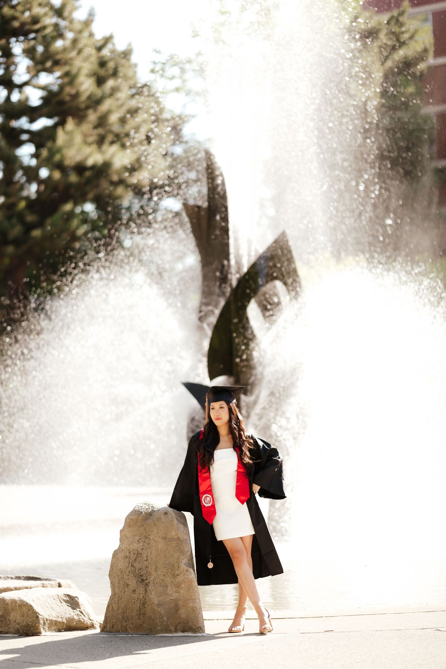 Graduate posing near a fountain spray wearing academic regalia and a black coat.
