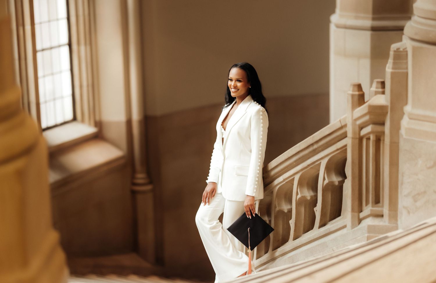 A woman in white business attire poses elegantly on a grand staircase in warm lighting.