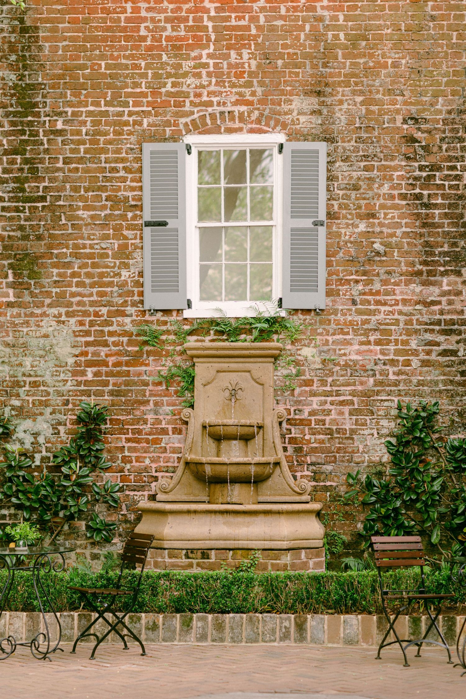 Historic brick wall with gray shuttered window and ornate stone fountain beneath.