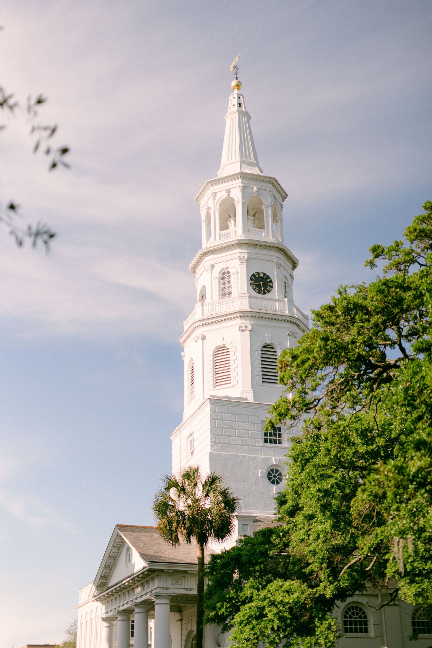 Majestic white church steeple with clock tower rises against blue sky surrounded by green trees.