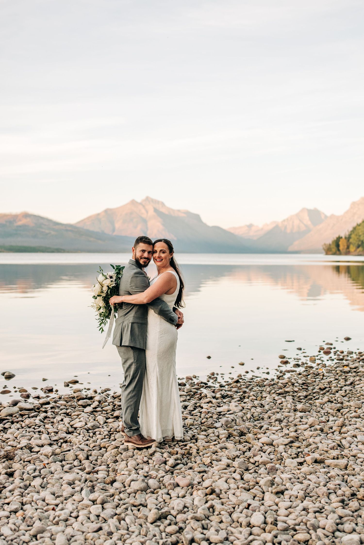 Wedding couple embracing on rocky beach shore with mountain backdrop at sunset.