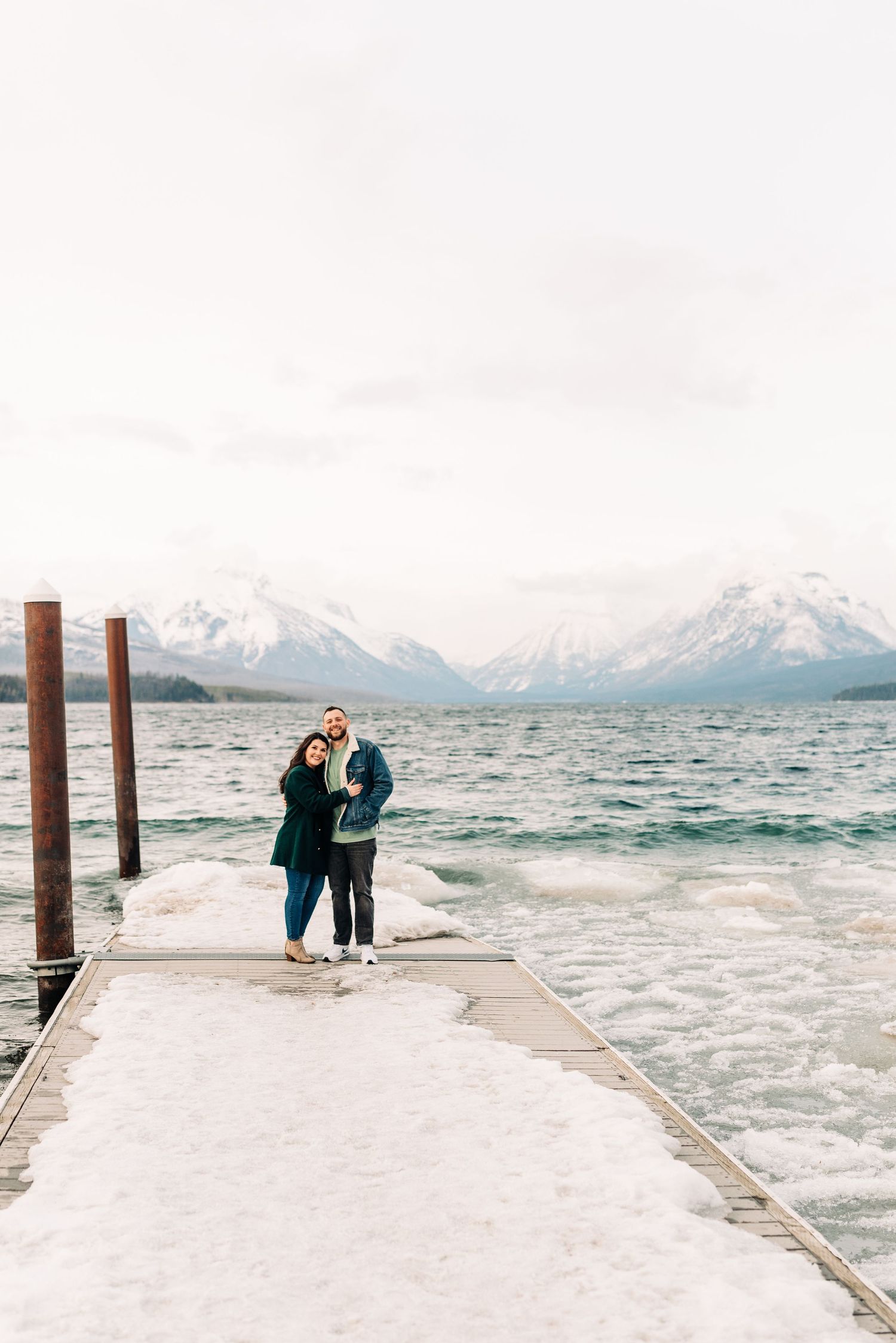 Couple standing on snowy dock with waves and mountain views at Lake McDonald.