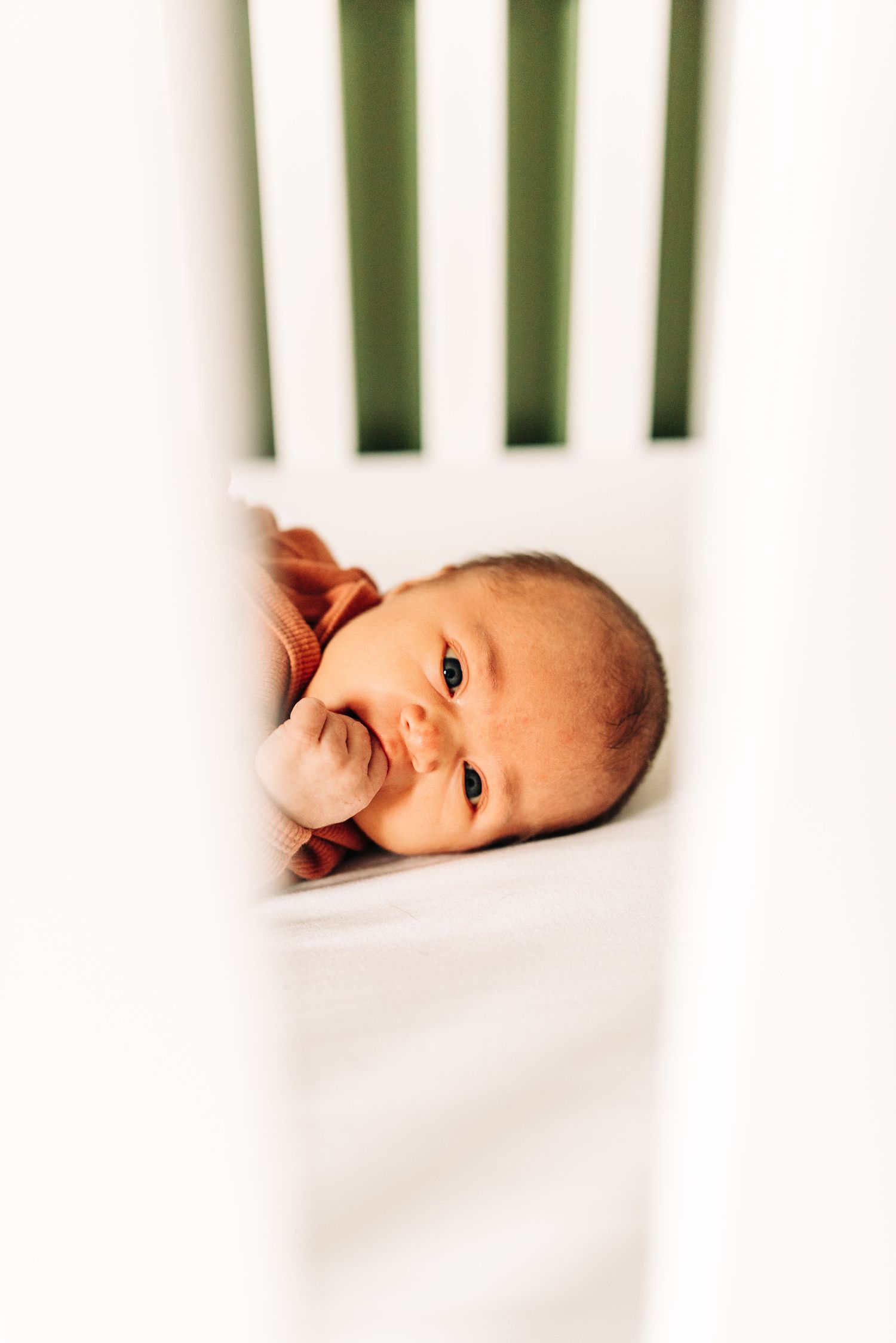 A close-up view through white crib slats shows a small infant lying on white bedding.