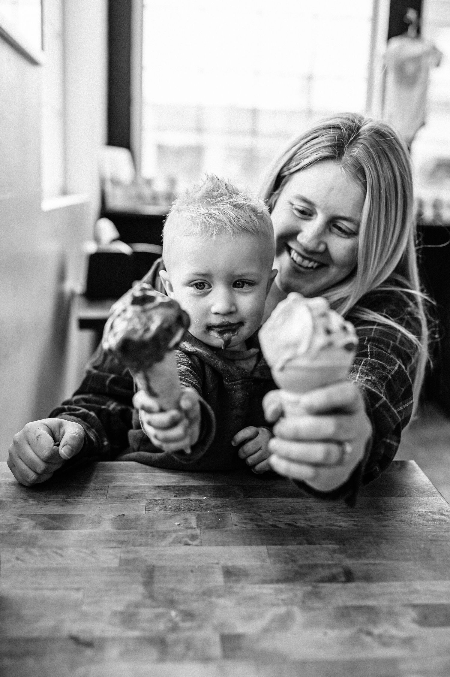 Mother and son sitting at a table together eating ice cream, both are holding their cones out towards the camera