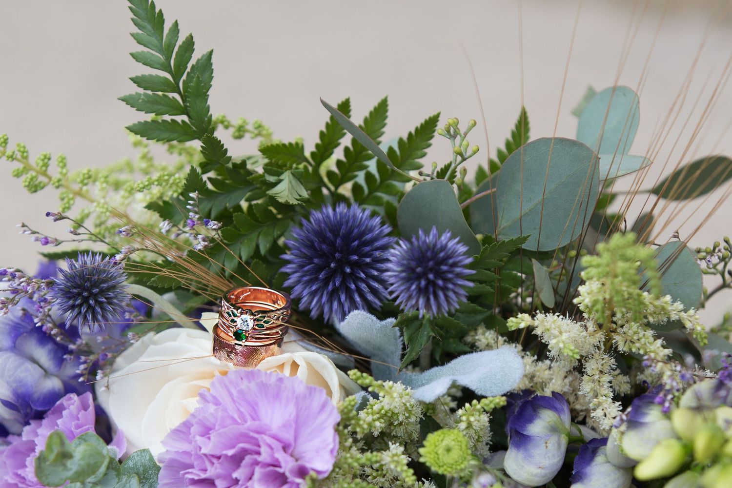 Groom's and bride's wedding and engagement rings sit stacked on a white rose in a white, purple,  and green flower bouquet.