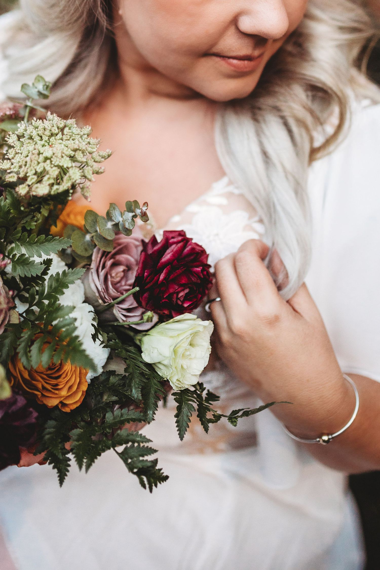 Bride in white dress holds a lush bouquet of burgundy, mauve, and yellow roses with greenery.