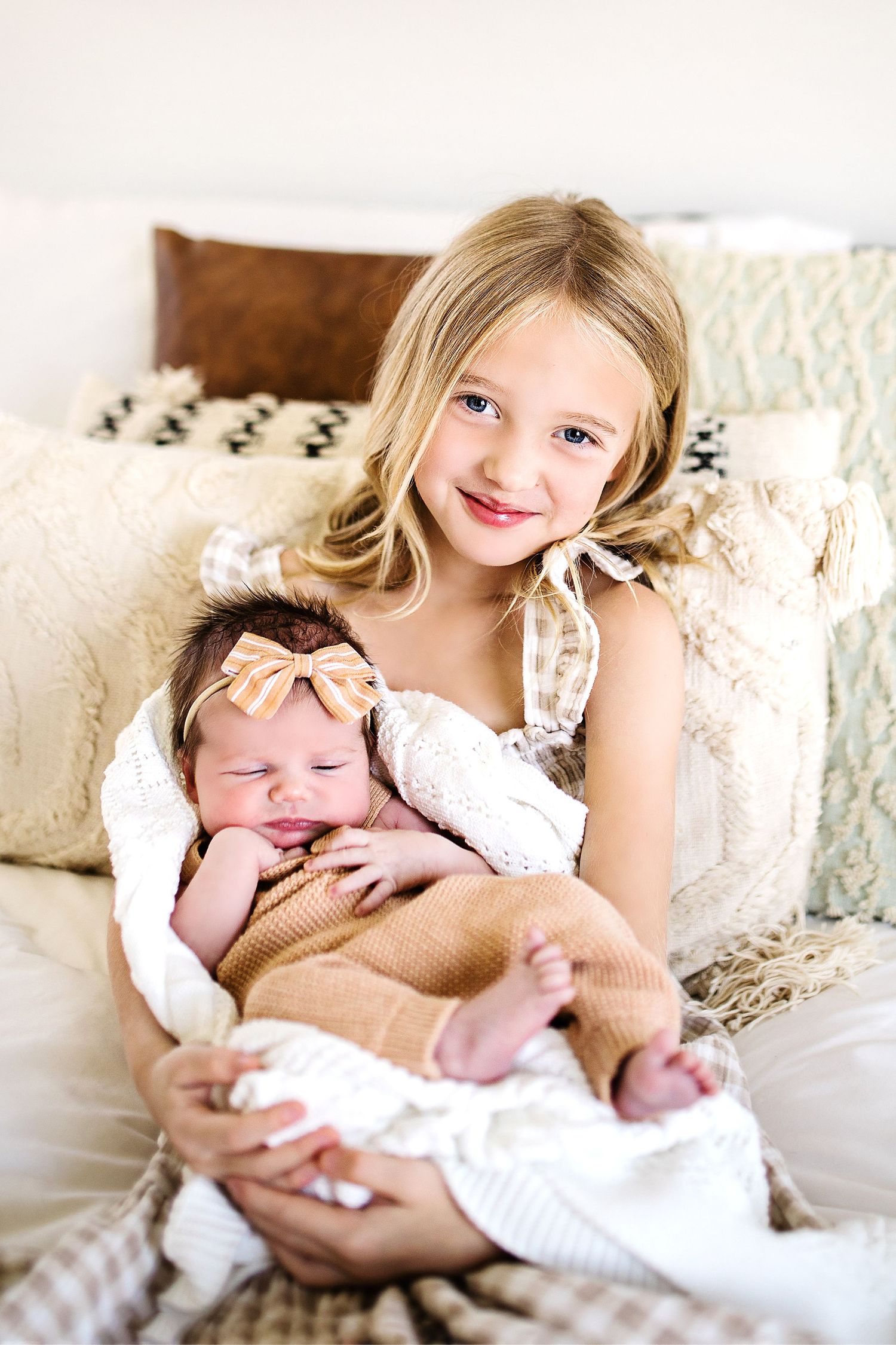Smiling blonde girl holds her newborn baby sibling on a cozy bed with decorative pillows.