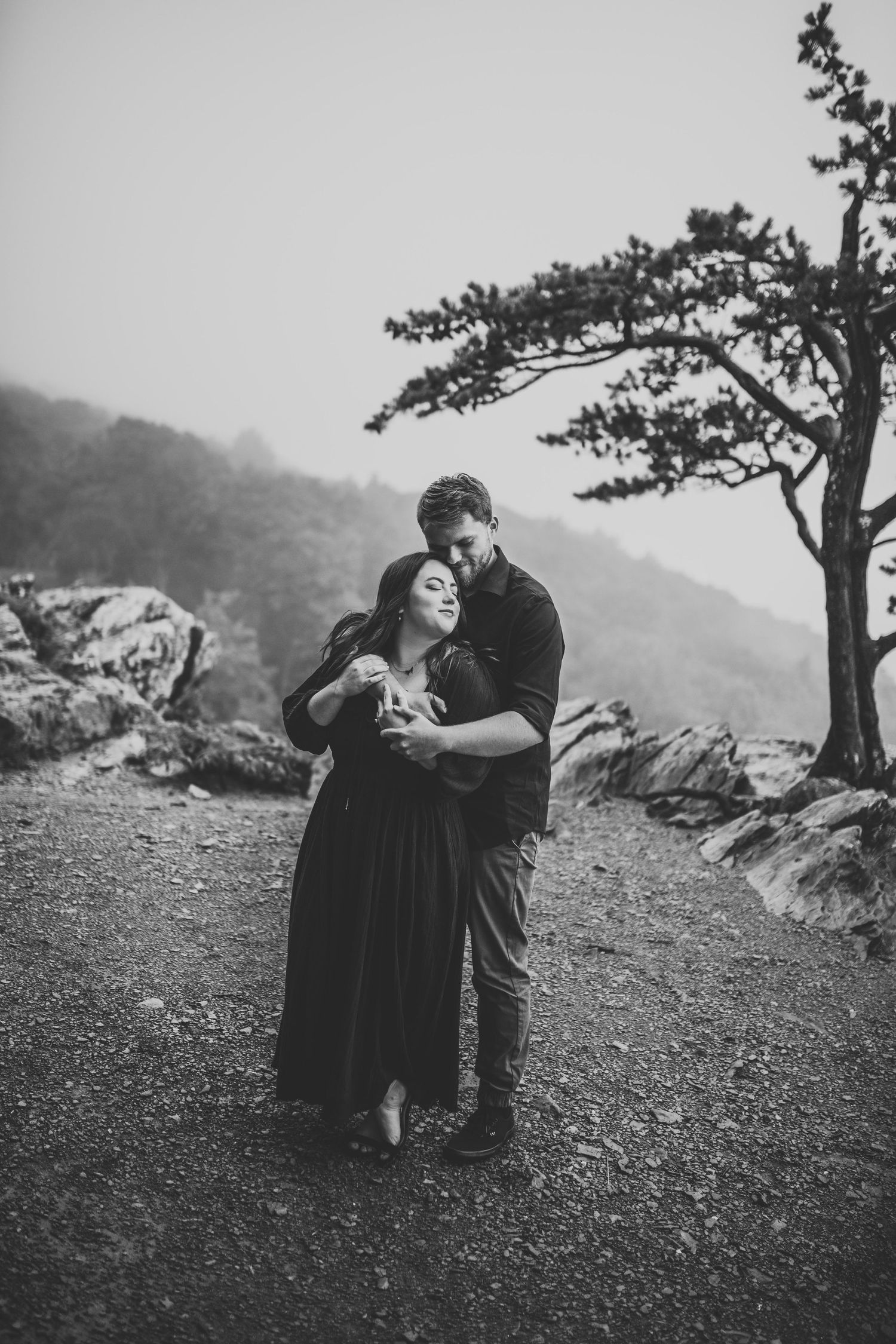 Black and white photo of couple holding each other on a misty mountain path with a tree.