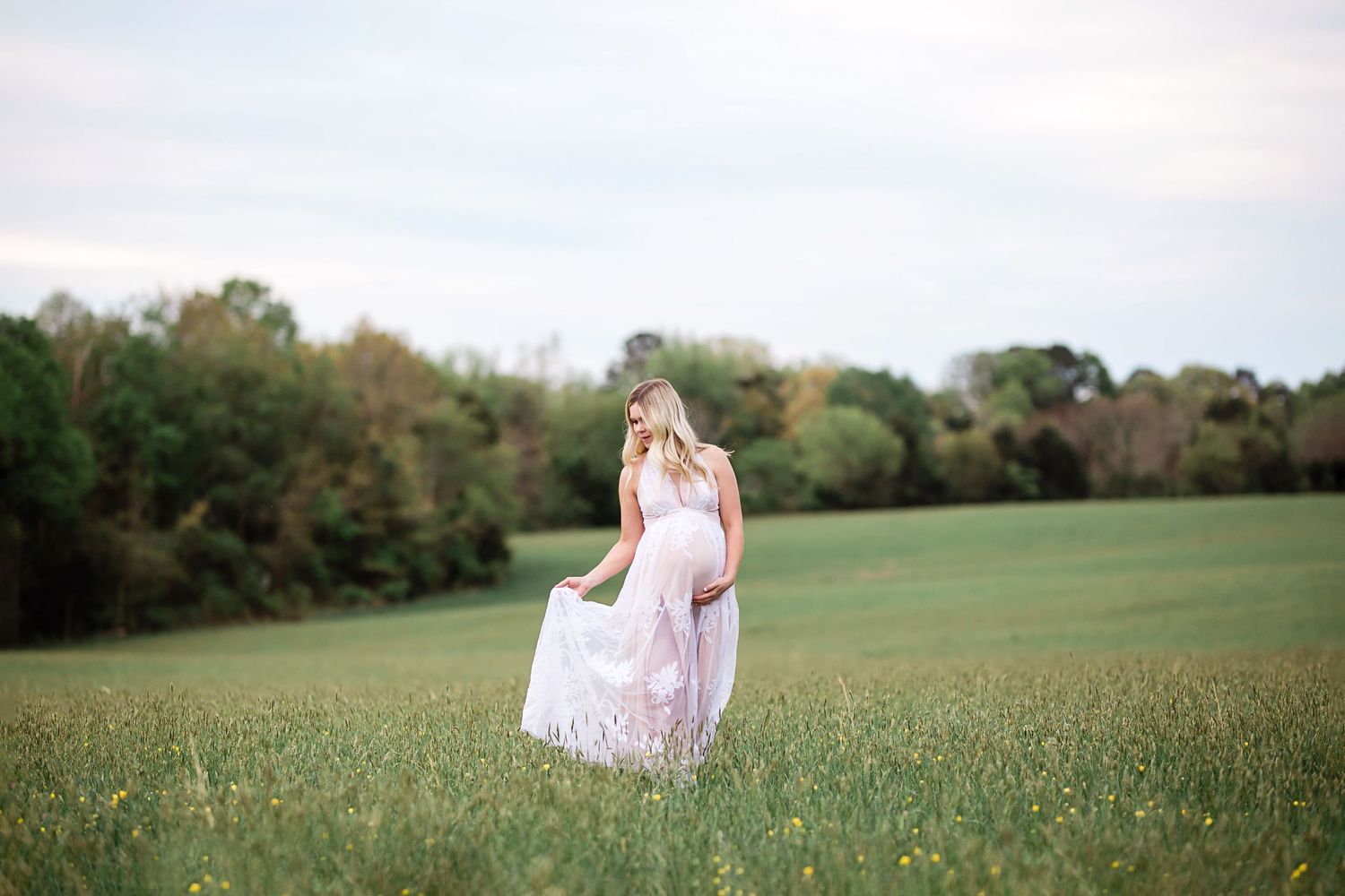 Pregnant woman in flowing pink gown walking through a green meadow with trees in the background.