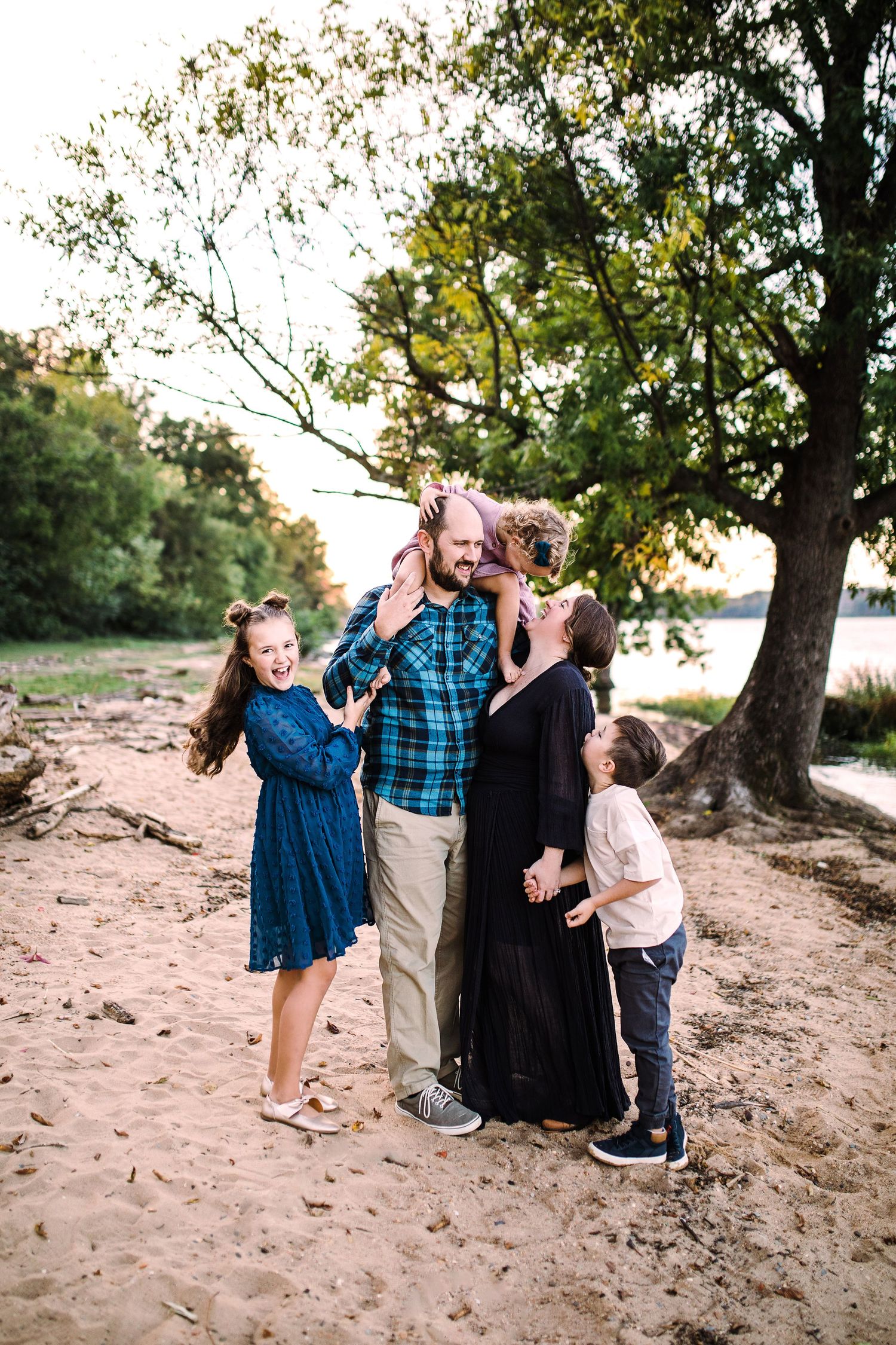Happy family of five laughing and embracing on a sandy riverbank surrounded by trees.