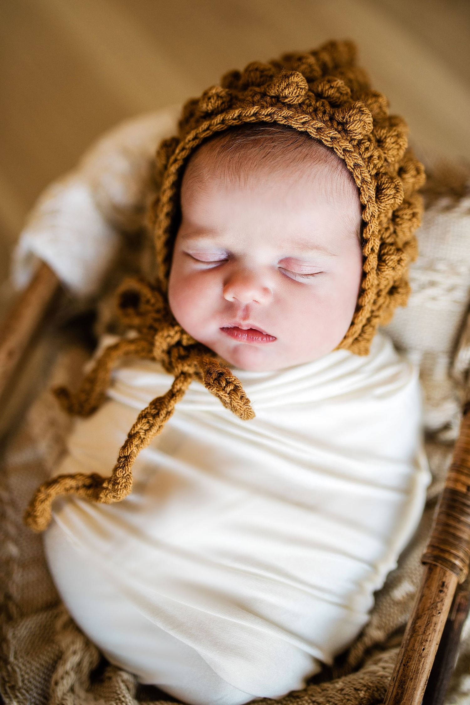 Sleeping newborn baby wrapped in white swaddle wearing a golden brown crocheted bonnet.