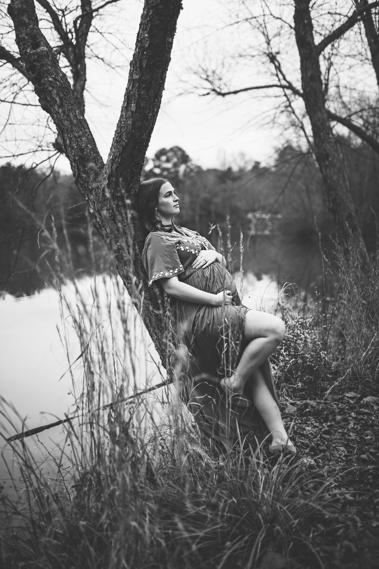 Black and white artistic photo of pregnant woman leaning against tree by a tranquil lake.