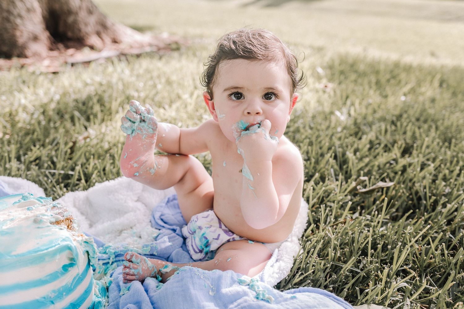 A baby in a light blue dress sits in a grassy field on a sunny summer day.  Pearland Family Photographer