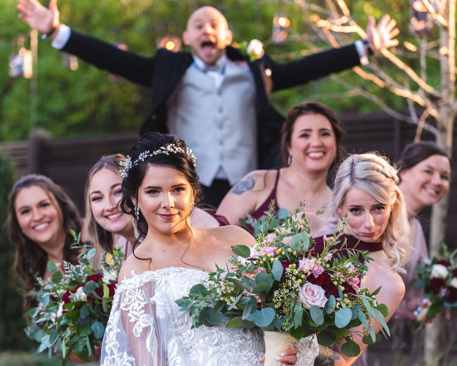 A bride with her bridal party, the groom is in the background jumping.