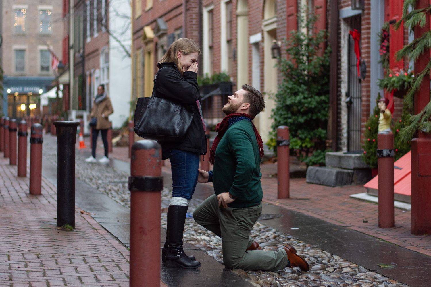 A man on his knees proposing to a woman standing next to him, she has her hands over her face.