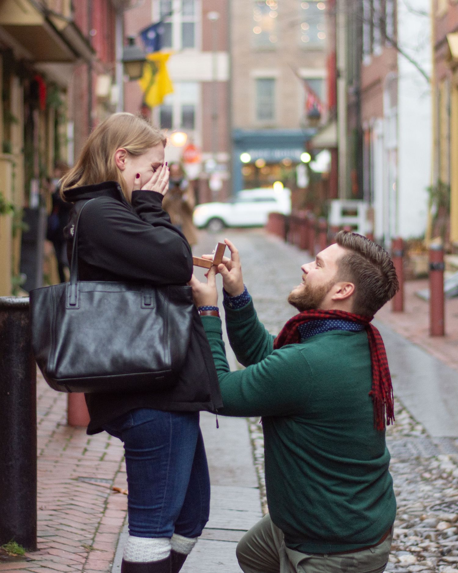 A man on his knees proposing to a woman standing next to him, she has her hands over her face.