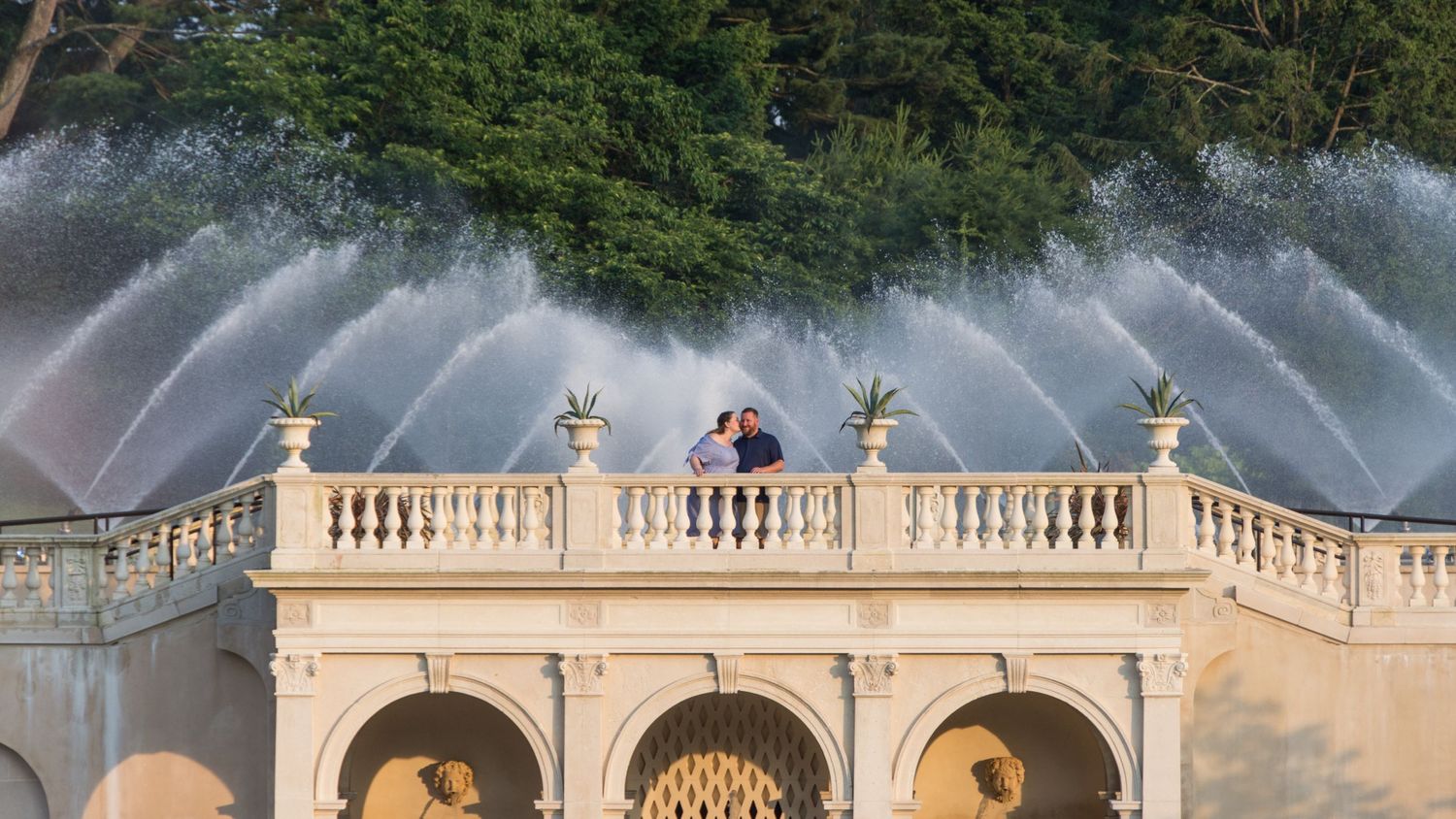 A newly engaged couple kissing on top of a bridge, water fountains are spraying behind them.