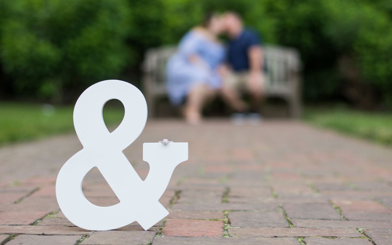 A newly engaged couple kissing, blurry in the background of an in-focus ampersand holding her ring.